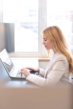 A businesswoman sits at a desk in a bright office, typing on a laptop. She is focused on her work, with natural light coming through the window, creating a professional and productive atmosphere.