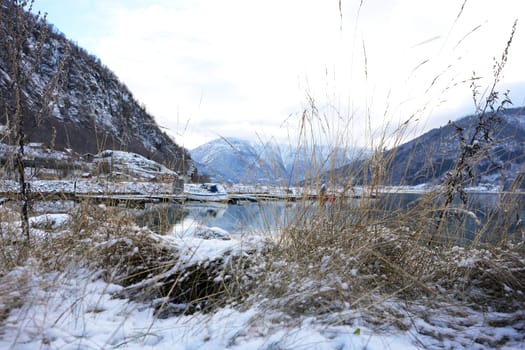 A scenic winter marina in Gaupne, Norway, with snow-covered mountains, calm waters, and boats docked along the fjord's edge. 