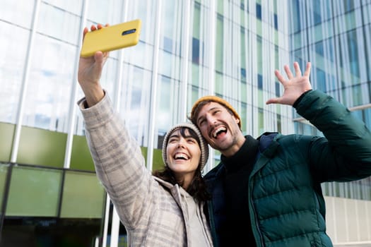 Cheerful man and woman taking a selfie with a yellow smartphone, waving and smiling in front of a modern office building