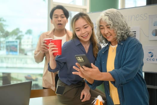 Group of cheerful colleagues enjoying drink and looking at smartphone during an office party.