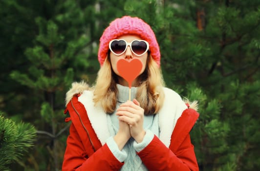Winter portrait of happy young woman having fun with sweet red heart shaped lollipop wearing pink hat, white glasses against Christmas tree in forest
