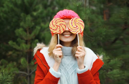 Winter portrait of happy funny smiling young woman having fun with sweet lollipop wearing hat against Christmas tree in forest