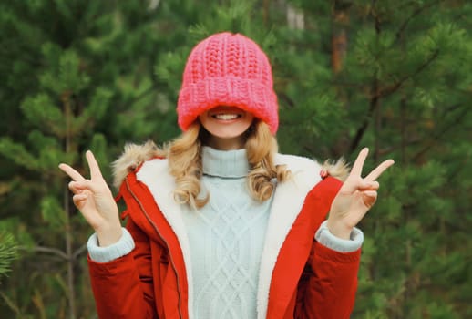 Happy smiling young woman having fun enjoys snow, warm weather in winter park against Christmas tree, joyful girl in hat standing in forest with snowflakes, festive background
