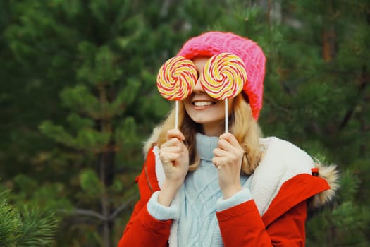Winter portrait of happy funny smiling young woman having fun with sweet lollipop wearing hat against Christmas tree in forest