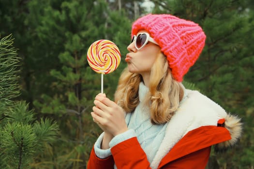 Winter portrait of happy funny smiling young woman having fun with sweet lollipop wearing hat against Christmas tree in forest