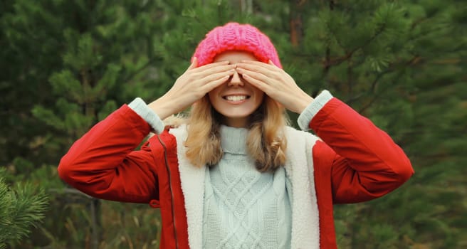 Happy smiling young woman having fun enjoys snow, warm weather in winter park against Christmas tree, joyful girl in hat standing in forest with snowflakes, festive background
