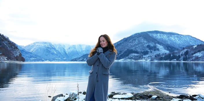 Cheerful woman enjoying the peaceful winter scenery by a snow-covered fjord in Gaupne, Norway, surrounded by mountains