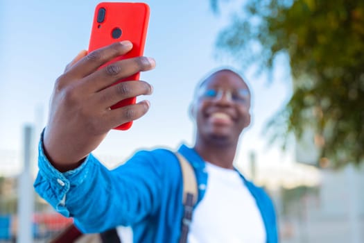 A young man smiles while taking a selfie outdoors with a red smartphone. He is dressed casually in a blue jacket and backpack. The background shows greenery and a bright, sunny atmosphere.