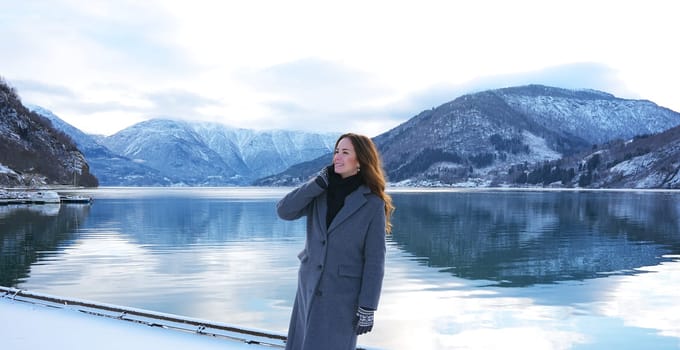 A cheerful woman in a coat and mittens smiles while admiring the tranquil snowy fjord and mountains in Gaupne, Norway