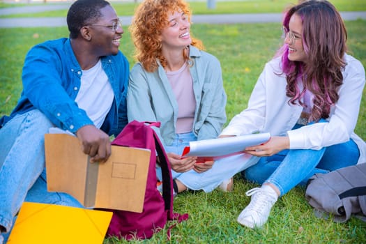 A diverse group of students sits on the grass outdoors, smiling and talking. They hold notebooks and are dressed casually, enjoying a moment of friendship and connection in a peaceful natural setting.