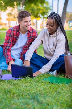 A young couple sits together on the grass outdoors, smiling and using a laptop. They are relaxed and enjoying their time, with bags and books around them, in a calm and bright natural setting.