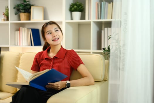 Charming young asian woman sitting on couch with book and looking out the window.