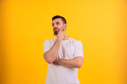 A young man is standing against a bright yellow background, looking up thoughtfully with a hand on his chin. He is in a casual t-shirt, appearing to be deep in thought or considering an idea.