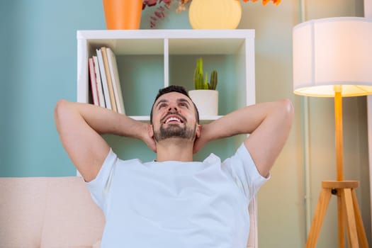 A man is relaxing on a sofa, smiling and looking up with his hands behind his head. He is in a modern, cozy living room with soft lighting, plants, and a comfortable atmosphere.