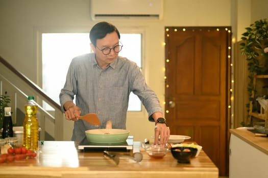 Happy middle aged man wearing glasses cooking on the counter in warm home kitchen.