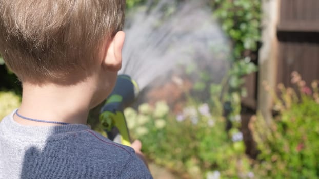 Funny little boy watering lawn plants in garden housing backyard. Adorable child playing with irrigation hose at hot sunny summer outdoors. Children help with housework. activity for kids. Childhood.