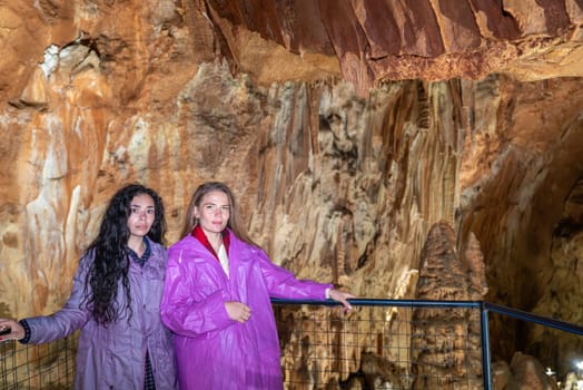 Two women standing in front of a cave with a railing. One of them is wearing a purple jacket