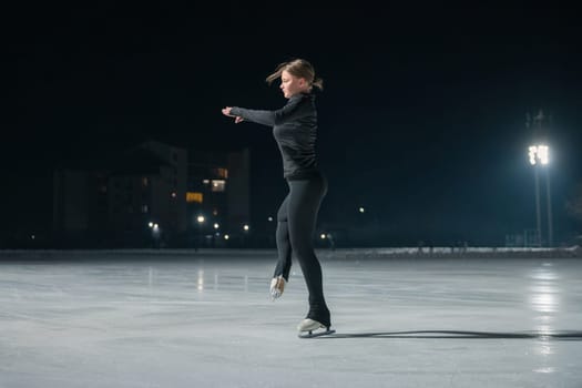A young woman in black athletic wear gracefully ice skates on a lit outdoor rink at night, surrounded by soft lighting.