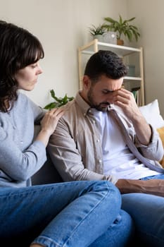 Vertical portrait of woman consoling sad husband sitting on the sofa at home living room. Lifestyle concept.