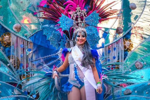 The image features a person dressed in a vibrant and elaborate costume, typically seen in carnivals or festive parades. The costume is adorned with a multitude of feathers and sequins in a dazzling array of colors, predominantly blue and pink tones. The person is also wearing a headpiece with intricate designs and decorations. They appear to be smiling and participating in a festive event, as indicated by a sash over their shoulder, which is often a sign of someone in a role of honor or as part of a pageant. Overall, the image captures a celebratory and colorful atmosphere.