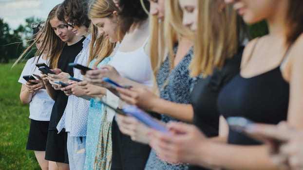 Group of teenage students are using smartphones outdoors in a park, standing in a line.