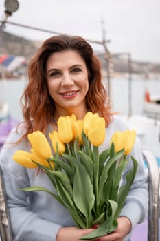 Woman holds yellow tulips in harbor with boats docked in the background., overcast day, yellow sweater, mountains.