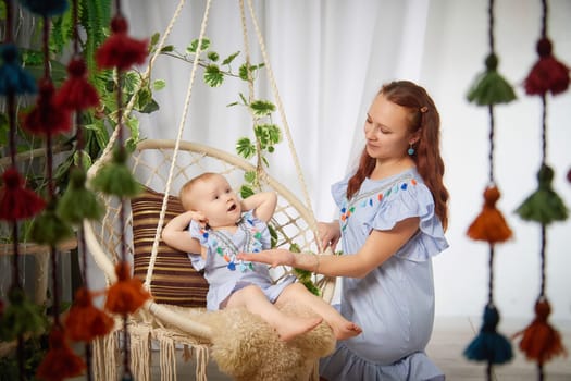 A mother and her daughter are sitting in hanging chair, the mother is smiling as the little girl reaches out to touch her