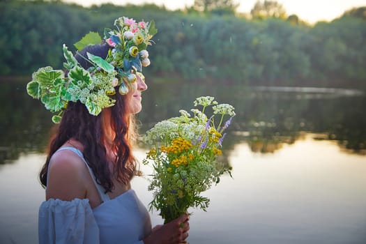 Slavic plump chubby girl in long white dress on the feast of Ivan Kupala with flowers and water in a river or lake on summer evening