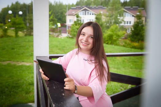 A woman smiles as she looks at her tablet, sitting on porch with a grassy view and houses in the background