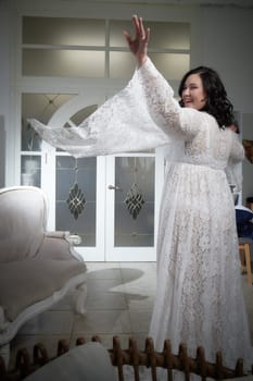 A plump woman spins gracefully in a white lace dress, embracing the moment in an elegant indoor setting filled with natural light.