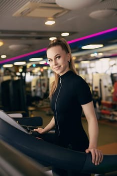 A woman in a fitted black outfit smiles confidently while using a treadmill in a contemporary gym. Natural light filters through large windows, creating an inviting atmosphere.