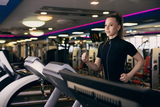A young woman is jogging on a treadmill in a well-equipped gym, surrounded by various exercise machines and bright lighting, showing focus and determination in her workout.