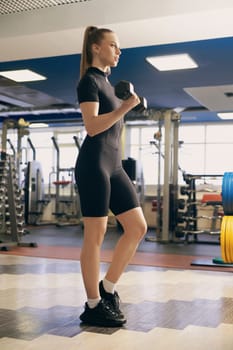 A young woman stands confidently in a gym, lifting dumbbells to enhance her strength. She wears a fitted athletic outfit and focuses on her workout routine in a bright, spacious environment.