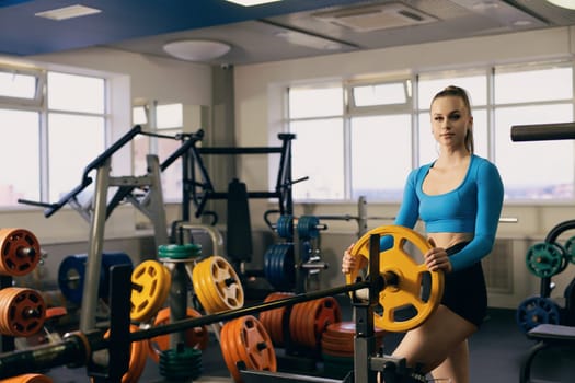 A young woman in a blue crop top prepares to lift a weight in a modern gym filled with colorful weight plates and exercise equipment, showcasing dedication to fitness and strength training.