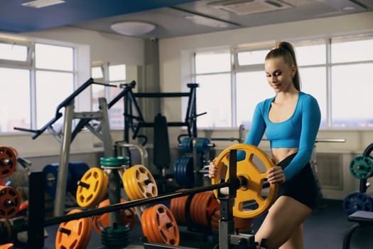 A young woman adjusts weights on a barbell in an equipped gym. The spacious area features colorful weight plates and large windows allowing natural light to flood in.
