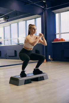 A woman in athletic wear completes squats on a step platform in a well-lit fitness studio. The environment is designed for exercise, with colorful weights visible in the background.