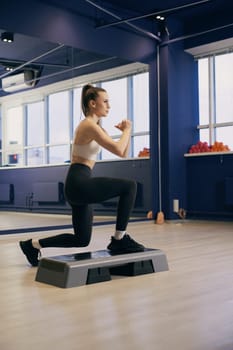 A young woman executes step lunges on an elevated platform in a bright fitness studio, focusing on her form. The space features large windows and fitness equipment in the background.