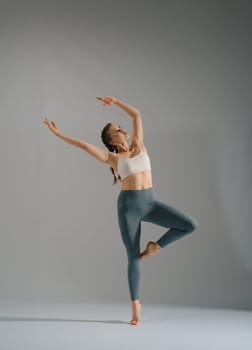 A dancer with flowing hair strikes a dynamic pose with one leg raised, surrounded by a neutral gray background that enhances her movement and elegance, showcasing her athleticism and artistry.