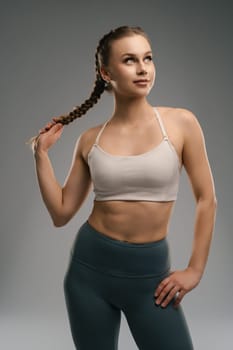 A young woman stands with a confident expression, wearing a sports bra and high-waisted leggings. She has a braided hairstyle and maintains an athletic pose, showcasing her fit physique.
