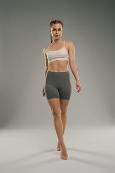 A fit model strides forward in a sports bra and shorts, showcasing her athletic physique. The neutral background highlights her powerful stance and confidence during the shoot.