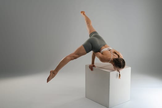 A dancer showcases a graceful pose while balancing on a white cube in a minimalist studio. The composition emphasizes movement and flexibility, drawing attention to the dancer's form.