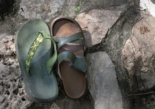 Saraburi, Thailand. Mar - 20, 2022 - Green beach flip flops for man and Brown summer slipper for woman are placed side by side on rocks by the waterfall with soft sunlight. Concept of Sometimes the long journey of life should rest, use it as your Wallpaper, Poster and Space for text. Selective focus.
