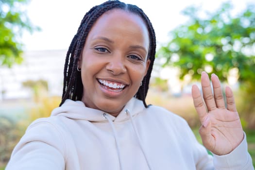 A smiling woman takes a selfie outdoors while waving with her hand. She is dressed in a light hoodie, enjoying the moment in a sunny environment with a bright and happy expression.
