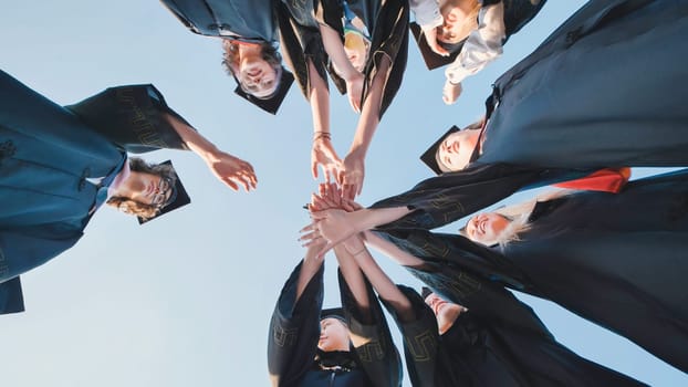 Group of young graduates wearing caps and gowns are joining their hands together in a circle, celebrating their achievement