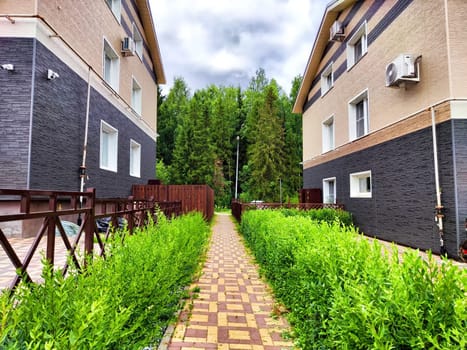 A pathway lined with greenery connects two modern residential buildings under a cloudy sky.