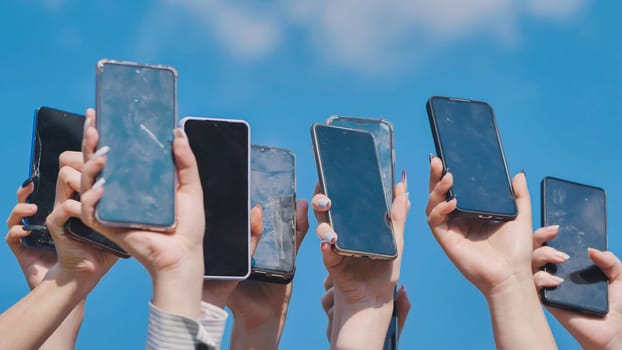 Many hands holding up smartphones with cracked and broken screens against a clear blue sky