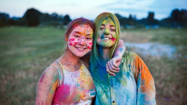 Two women covered with colorful holi powder are laughing and enjoying the event on a field