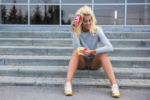 Young woman sitting on stone steps, holding a soda can and snack, enjoying a sunny day in a park setting.
