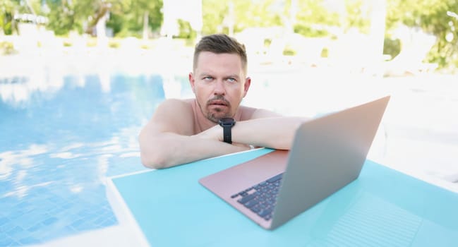 Annoyed man working on a laptop from the pool, close-up. Unexpected remote work while on vacation, distraction from rest