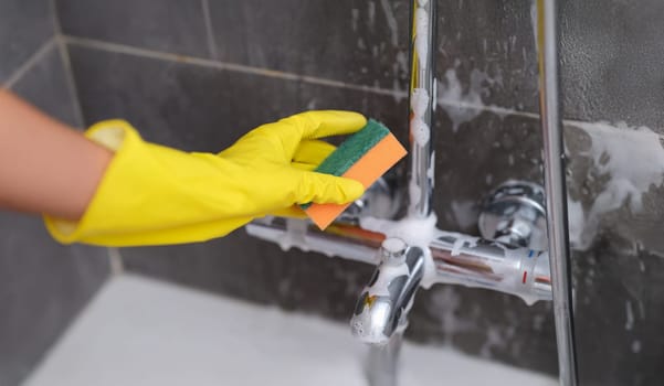 A hand in a glove washes the mixer with a sponge in the bathroom, close-up. Detergent for plumbing, disinfection